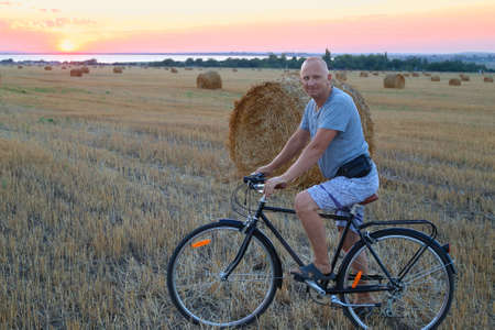 An adult man on a bicycle on a mowed field next to a straw walker at sunset. Sports and holiday activities in the countryside.の写真素材