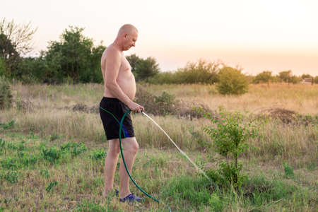 A mature man stripped to the waist is watering seedlings of fruit trees from a rubber hose. Summer...
