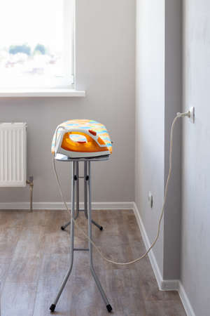 The electric iron is plugged into the socket on the ironing board in the room against the background of the window. Iron clothes.の写真素材