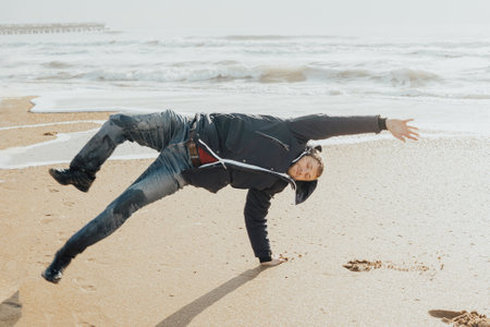 A young man in a jacket does somersaults on his hands on a sandy shore on a winter day. Travel and tourism.の写真素材