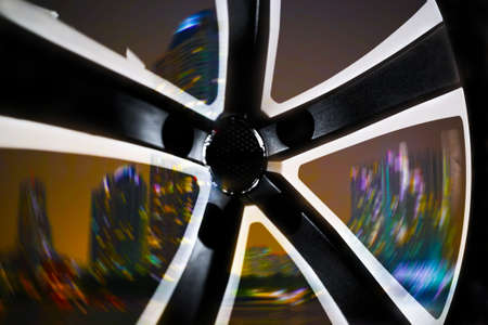 Black alloy wheel of a premium car in motion, close-up. Blurred night city in the background.