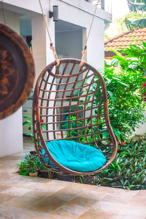 Wicker hanging cozy chair with a pillow on the site near the house, next to the tropical garden....