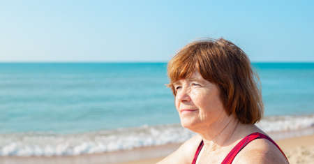 Portrait of a smiling elderly woman sitting on the seashore on a sunny day. Summer pastime in...