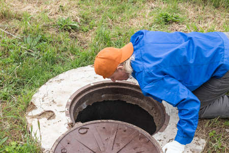 A working plumber bent over a water well to inspect pipes and a meter. Rural water supply, repair and maintenance.の写真素材