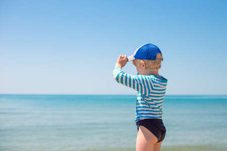 A little boy in a striped T-shirt and a blue cap stands on the seashore and looks into the distance. Happy summer days. copyspace.の写真素材
