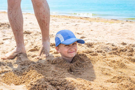 The boys head sticks out of the sand, next to his fathers legs. Fun fun at sea with family during summer holidays.の写真素材