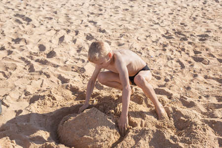 A fair-haired boy builds a sand tower on the beach on a summer sunny day. happy holidays.の写真素材