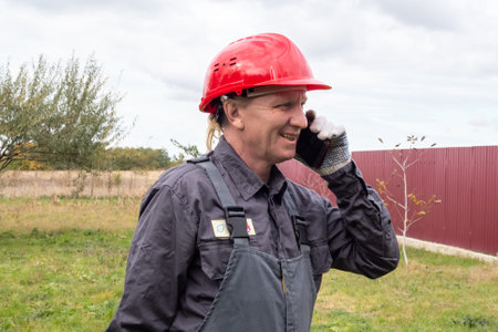 An adult male worker in overalls and a hard hat is talking on the phone on the street and smiling.