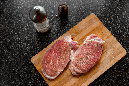 Pieces of raw meat sprinkled with salt and spices on a cutting board, on a black tabletop. Cooking pork steaks chops.の写真素材