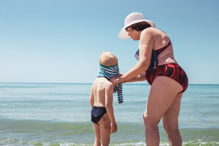 Grandmother dresses her grandson on the seashore, after swimming. Summer vacation on the sea coast with the whole family.の写真素材