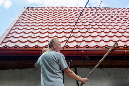 A mature man cleans the gutter of a drainpipe on the roof of his house from autumn leaves and debris.の写真素材