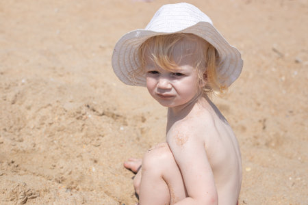 A little girl in a white hat plays in the sand, squinting in the sun.の写真素材