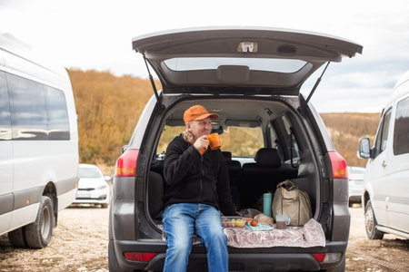 Caucasian man sits in the trunk of a car and drinks coffee. Outdoor picnic. Road trip.の写真素材