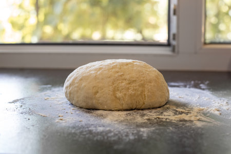 A lump of dough on a black table with scattered flour against the background of an open window....