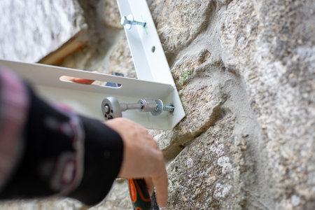 A person uses a ratchet wrench with a socket to screw a screw into a metal bracket to install an air conditioner on the stone wall of a rural home.の写真素材