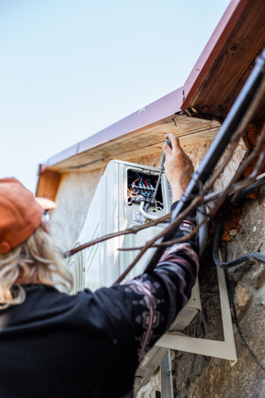 A man mounts an outdoor air conditioner unit on the wall of a stone farmhouse, installing electrical wiring and connecting a signal cable. DIY installation.の写真素材