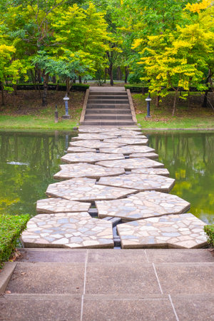 Large slab stone pavement leading across a pond to steps with green and yellow trees in the background in Chatuchak Park, Bangkok.の写真素材