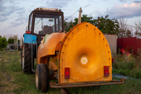 Blue tractor and orange agricultural sprayer on grassy field. Farm machinery for agricultural work.