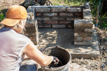 Person in cap mixing cement in bucket while building brick structure of outdoor barbecue.
