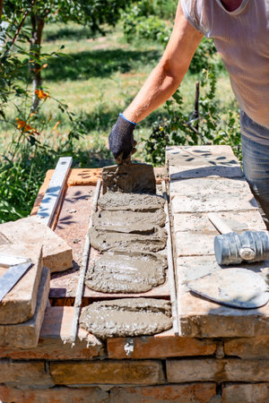 A worker applies mortar with a trowel and lays bricks, continuing the construction of a barbecue.