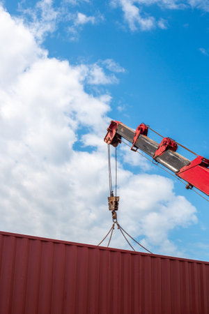 Red crane lifting red cargo container against sky and clouds.