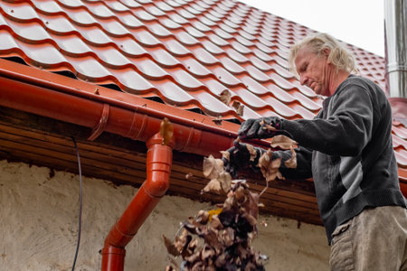 A man on a ladder clears leaves from the gutter of a house with a red tile roof.の写真素材