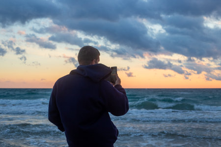 A man takes a photo of a golden sunset over the sea with his phone.