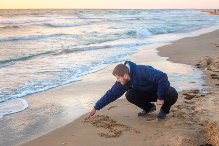 A man kneels on the beach, drawing with his finger in the sand next to gentle waves.の写真素材