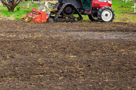 A small red tractor tills the soil in a field, preparing it for planting.の写真素材