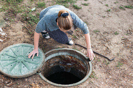 A woman peers into an open sewer manhole, inspecting the drainage system and septic tank water level.の写真素材