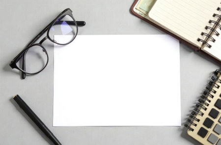 An overhead view of a wooden table with a blank white sheet of paper, surrounded by a calculator, glasses, pen, and notepad. Space for text or design.の素材