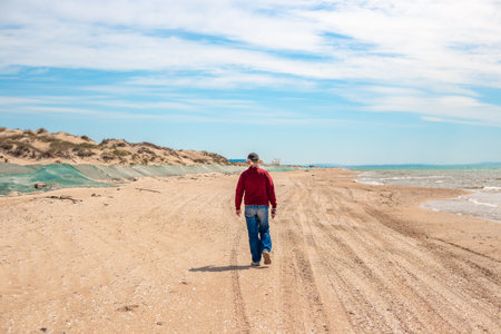 A man walks along a sandy beach on a sunny day. Along the sea, a sand embankment is covered with a net.の写真素材