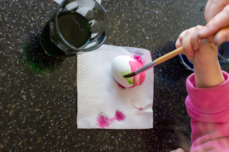 An adult and child carefully paint a white Easter egg with a brush, creating pink stripes for a...