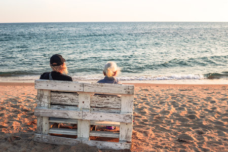 Two elderly people sit on a rustic wooden bench on a sandy beach, gazing at the calm ocean under a warm, clear sky.の写真素材