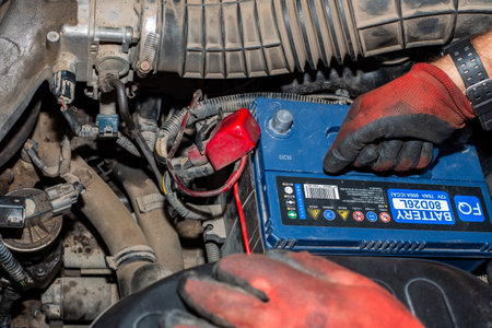 A mechanic lifts a blue car battery by the handle during battery maintenance and replacement. Anapa, Russia, May 20, 2023.のeditorial素材