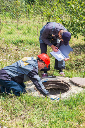 Male workers in overalls and holding a folder in their hands inspect a water well, checking the...