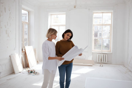 Two female interior designers discuss a layout while standing in a bright, white, empty room undergoing renovation.の素材