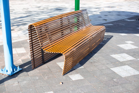 A modern wooden bench with slats sits on a paved public square in bright sunlight next to colorful...