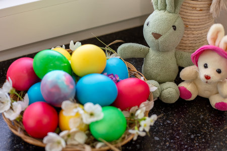 A straw basket filled with colorful Easter eggs sits next to plush rabbit toys. holiday decoration,...