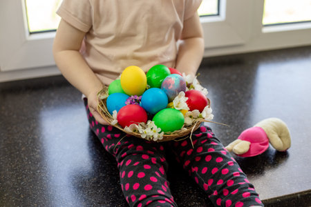 A child sits on a windowsill holding a basket of brightly colored Easter eggs. holiday tradition,...