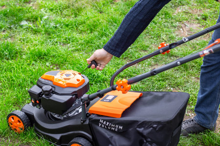 A man pulls the starter cord of an orange lawn mower on green grass. garden maintenance, lawn care,...