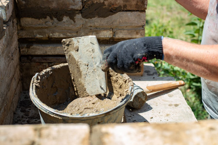 A hand in a black glove uses a trowel to take mortar from a bucket for garden construction. masonry work, building process, diy project.の写真素材