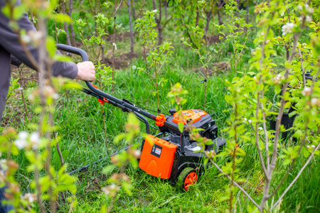 A man pushes an orange lawn mower through tall green grass and small trees in a garden. yard...