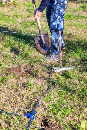 A man in camouflage pants digs into the earth with a shovel in a field. landscape work, gardening activity, soil digging.の写真素材