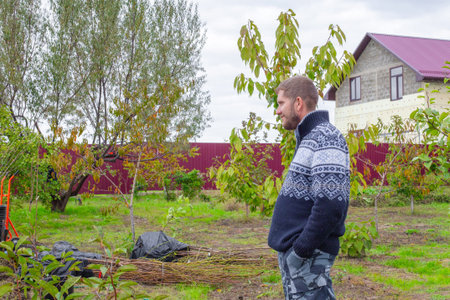 A man stands in a green field with young trees, looking left, with a house in the background. Gardening, rural lifestyle, plant care.の写真素材