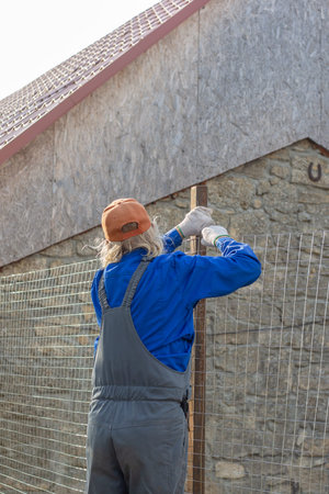 A worker in a blue jacket and orange cap installs a wire fence next to a stone building outdoors. fence installation, building work, home project.の写真素材