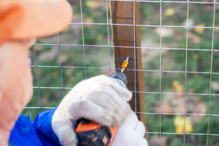 A worker in an orange cap uses a cordless drill to fasten wire mesh to a fence post. fence building, manual labor, power tool.の写真素材