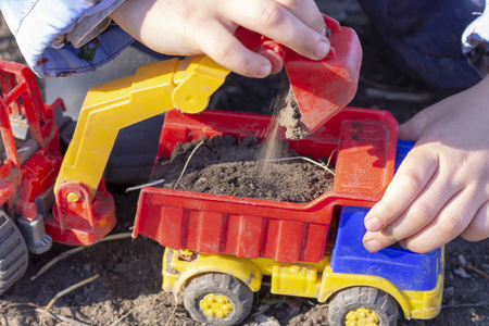 the child is playing on the street with sand; he loads the excavator bucket with earth into the dump truck toyの写真素材