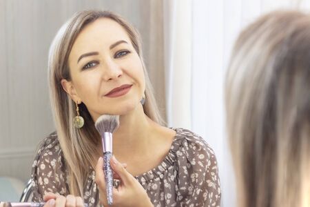 aged woman puts on her makeup. looking in the mirror. to myself a makeup artist. applying powder on the face with a large brushの写真素材