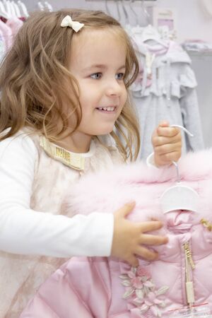 shopping. vertical photo. discounts. little girl shopaholic. girl delighted with beautiful jackets, chooses warm clothes. shopping center, shopping. emotionsの写真素材